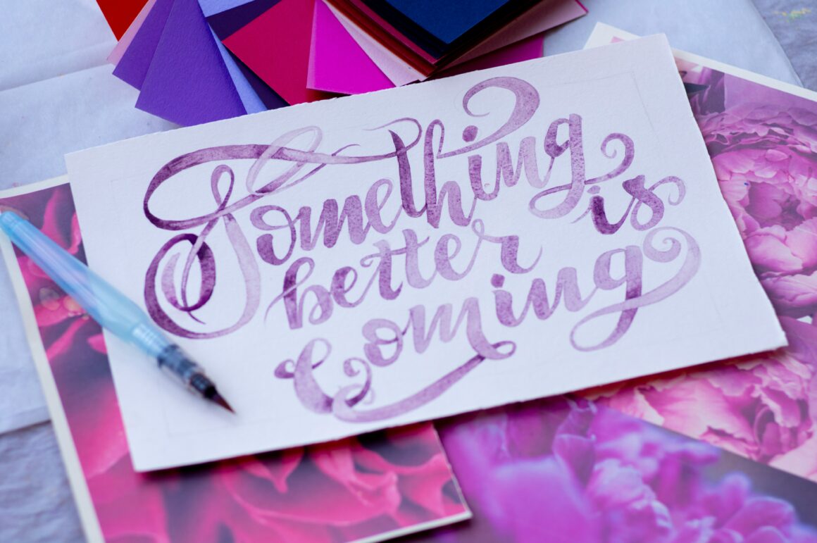 an artists desk is seen, on it someone has painted on white paper 'something better is coming' in purple writing to represent Affirmations for the Illness Journey