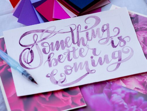 an artists desk is seen, on it someone has painted on white paper 'something better is coming' in purple writing to represent Affirmations for the Illness Journey