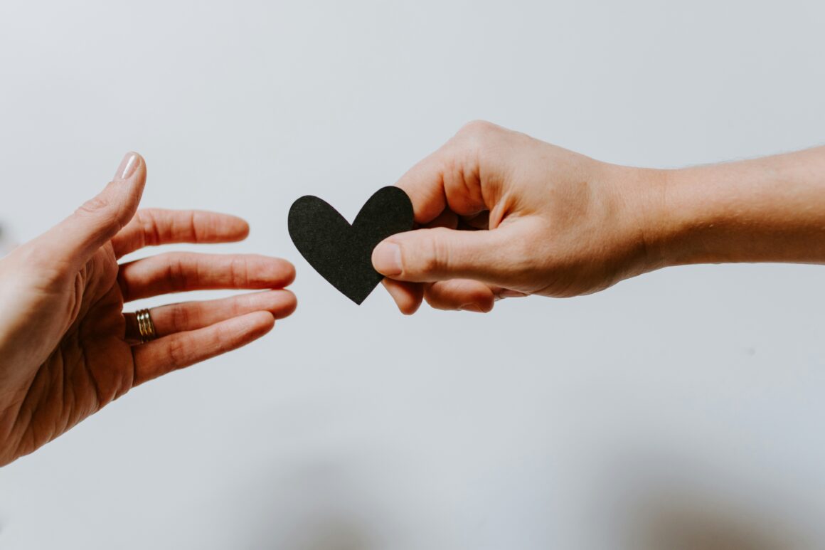 two hands are seen reaching out from either side of the picture. The one on the right is passing a small black heart this is against a white background.