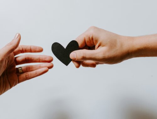two hands are seen reaching out from either side of the picture. The one on the right is passing a small black heart this is against a white background.