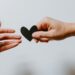two hands are seen reaching out from either side of the picture. The one on the right is passing a small black heart this is against a white background.