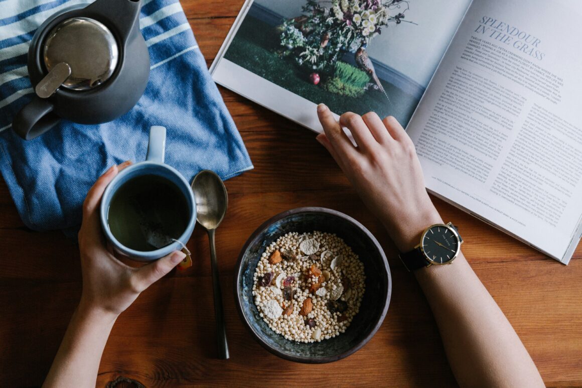 a person's arms are seen one os holding a cup of coffee, and the other flicking through a magazine on a brown table. There is also a coffee pot and a bowl of museli on the table to represent youtube creator wellbeing