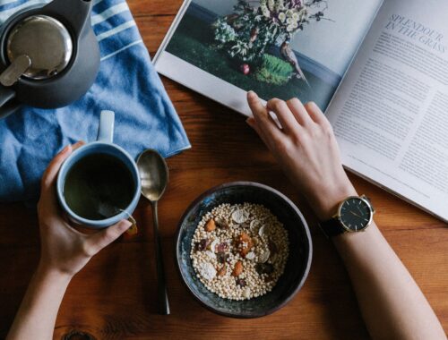 a person's arms are seen one os holding a cup of coffee, and the other flicking through a magazine on a brown table. There is also a coffee pot and a bowl of museli on the table to represent youtube creator wellbeing