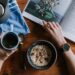 a person's arms are seen one os holding a cup of coffee, and the other flicking through a magazine on a brown table. There is also a coffee pot and a bowl of museli on the table to represent youtube creator wellbeing