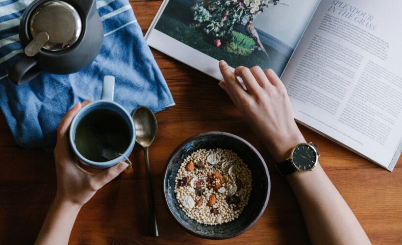 a person's arms are seen one os holding a cup of coffee, and the other flicking through a magazine on a brown table. There is also a coffee pot and a bowl of museli on the table to represent youtube creator wellbeing