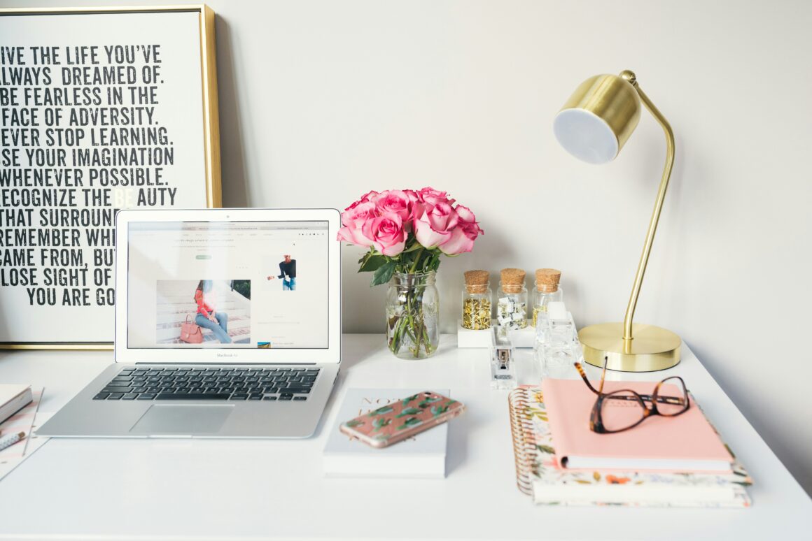 A white desk is seen, with a gold lamp, notebooks, glasses and a computer, beside them is a picture with inspirational quotes on it