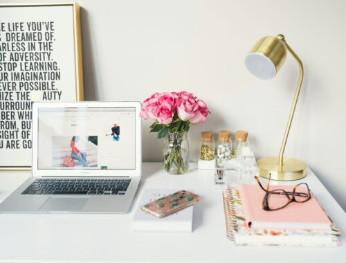 A white desk is seen, with a gold lamp, notebooks, glasses and a computer, beside them is a picture with inspirational quotes on it
