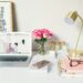 A white desk is seen, with a gold lamp, notebooks, glasses and a computer, beside them is a picture with inspirational quotes on it