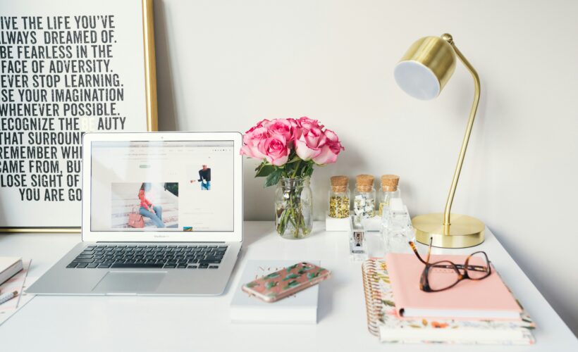 A white desk is seen, with a gold lamp, notebooks, glasses and a computer, beside them is a picture with inspirational quotes on it