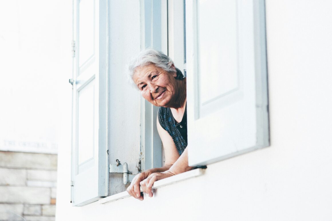 An older lady is looking out of a window at the camera