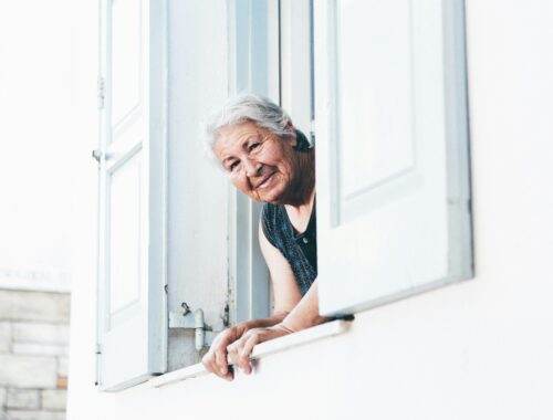 An older lady is looking out of a window at the camera