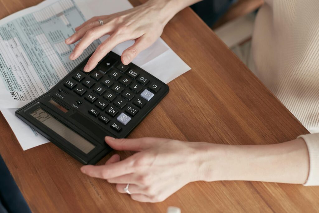 Two hands are seen working on a calculator on a brown desk. Under the calculator is a spreadsheet