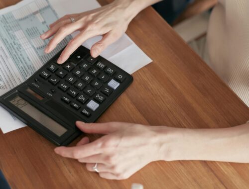 Two hands are seen working on a calculator on a brown desk. Under the calculator is a spreadsheet