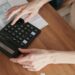 Two hands are seen working on a calculator on a brown desk. Under the calculator is a spreadsheet