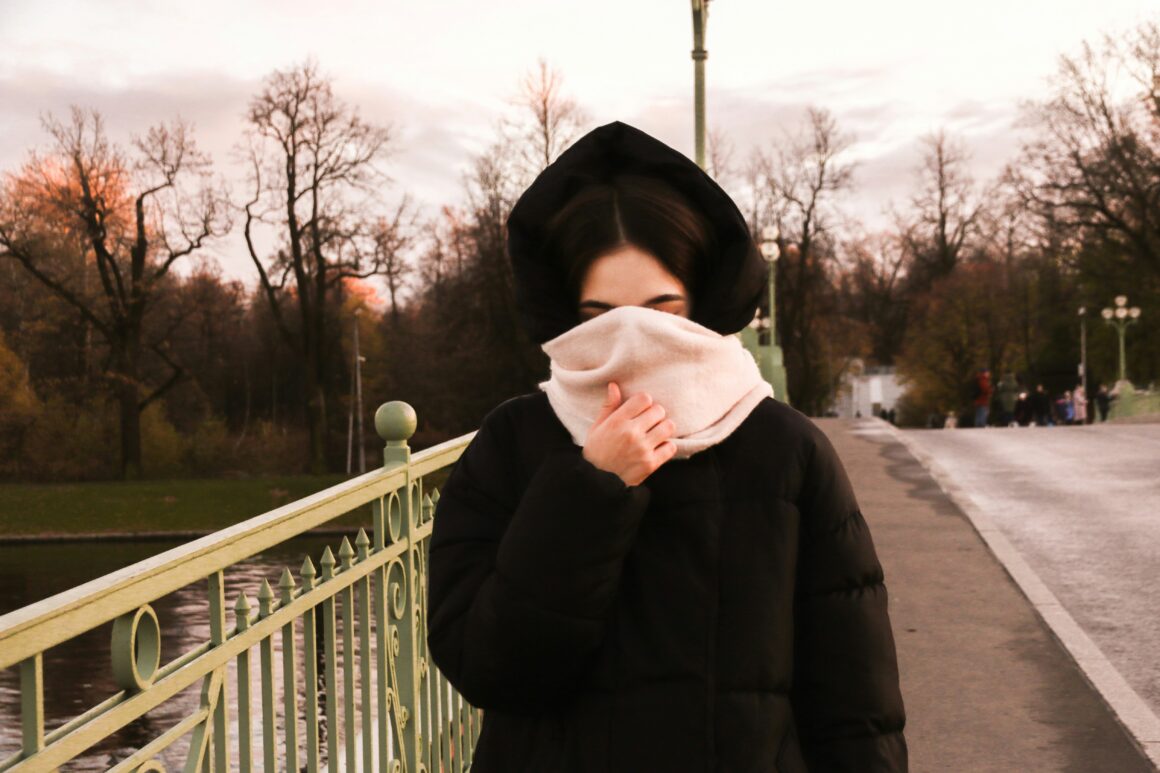 A woman in seen on a bridge she has a black coat on and her hood is up she has a thick pink scarf covering her face to represent Living Beyond the Visible: My Life with Invisible Disabilities