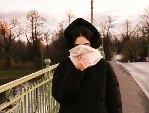 A woman in seen on a bridge she has a black coat on and her hood is up she has a thick pink scarf covering her face to represent Living Beyond the Visible: My Life with Invisible Disabilities