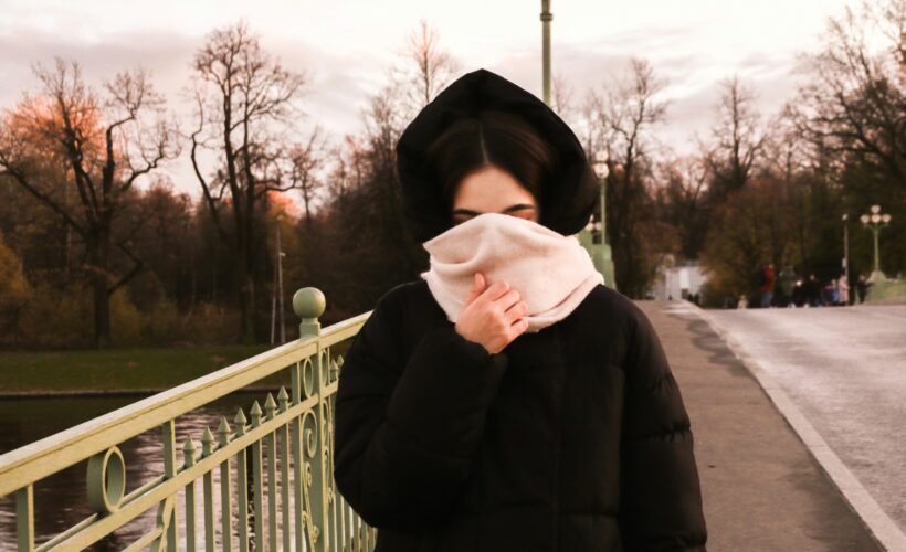 A woman in seen on a bridge she has a black coat on and her hood is up she has a thick pink scarf covering her face to represent Living Beyond the Visible: My Life with Invisible Disabilities