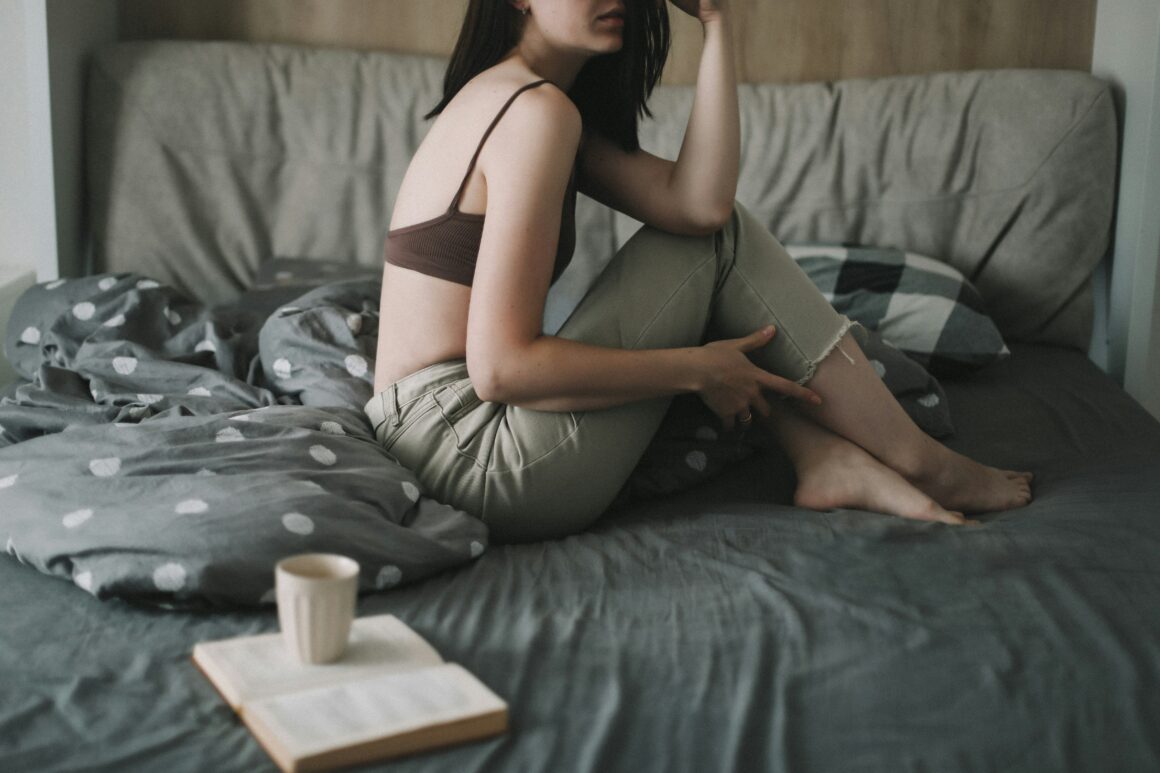 A woman in seen sitting on a bed, the bedding is grey and beside her is an open book with a mug of coffee on top