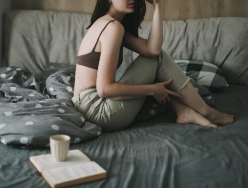 A woman in seen sitting on a bed, the bedding is grey and beside her is an open book with a mug of coffee on top