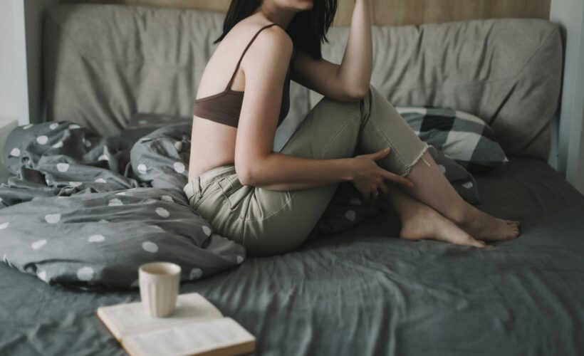 A woman in seen sitting on a bed, the bedding is grey and beside her is an open book with a mug of coffee on top