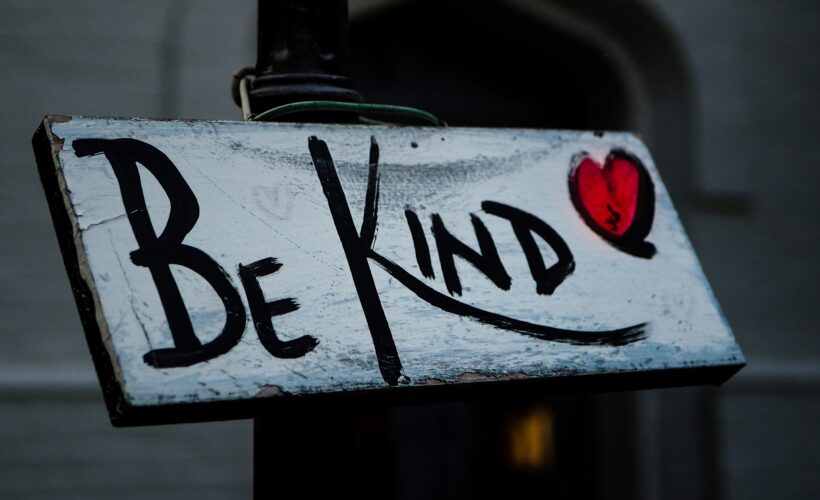 A sign is seen, painted white with a red heart and the writing Be Kind written in black to represent accessible ways to spread kindness