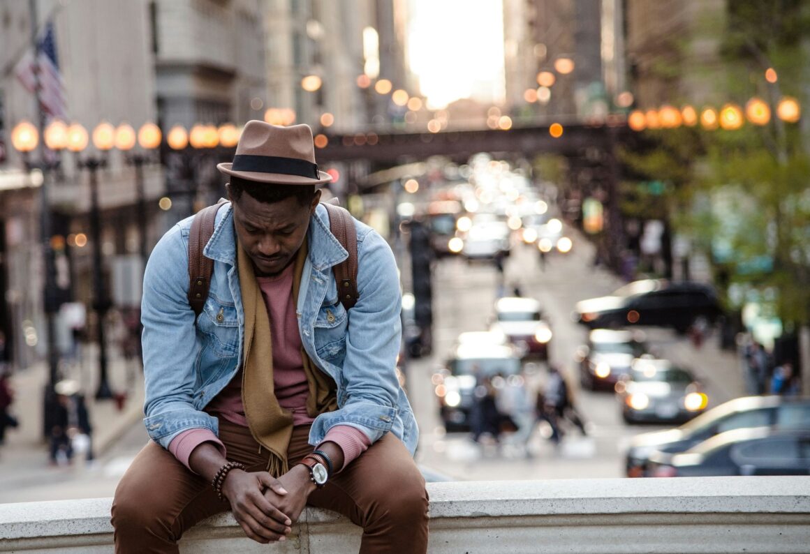 A man is sat on a low wall, behind him is a busy street, he is wearing a brown outfit under a blue denim jacket and a brown trilby on his head. He is looking down seemingly depressed and struggling to represent Managing Stress as a Disabled Carer