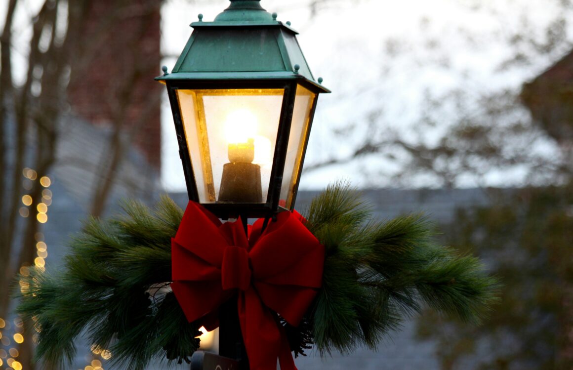 An old fashioned style lamppost is seen, close in on the lit part, below this section is tied a large red bow and fir brances, behind can be seen christmas lights out of focus