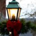 An old fashioned style lamppost is seen, close in on the lit part, below this section is tied a large red bow and fir brances, behind can be seen christmas lights out of focus