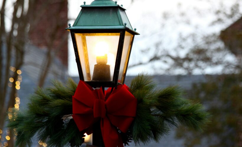 An old fashioned style lamppost is seen, close in on the lit part, below this section is tied a large red bow and fir brances, behind can be seen christmas lights out of focus