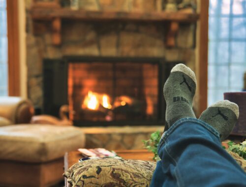 We see someone with cosy socks, their feet up in front of a roaring fire it is a cosy scene to represent How Late Winter Affects My Mental Health