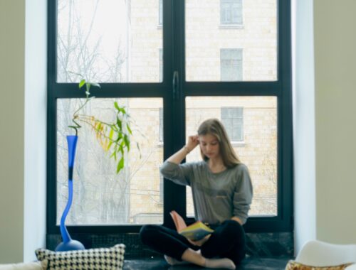 A woman is seen sitting in a window reading, beside her is a strangely shaped tall vase in blue she is pushing her hair back and looks tired.