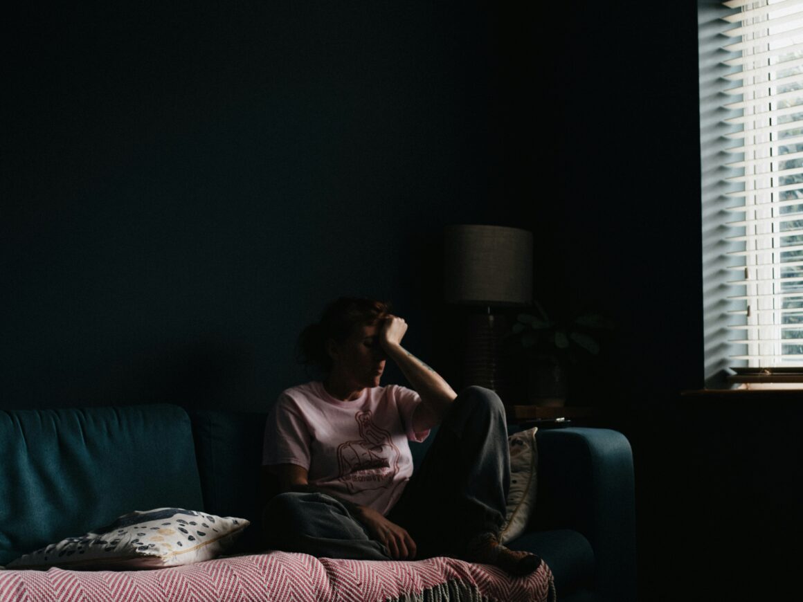 A person is seen sitting on a sofa near a window, they are curled up with their head in their hands and they are in low light to represent Wanting to Feel Seen When You’re Chronically Ill