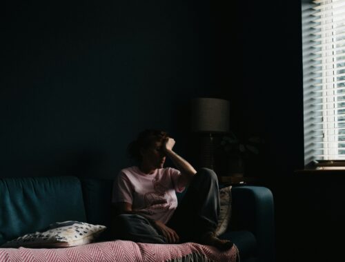 A person is seen sitting on a sofa near a window, they are curled up with their head in their hands and they are in low light to represent Wanting to Feel Seen When You’re Chronically Ill