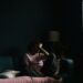A person is seen sitting on a sofa near a window, they are curled up with their head in their hands and they are in low light to represent Wanting to Feel Seen When You’re Chronically Ill