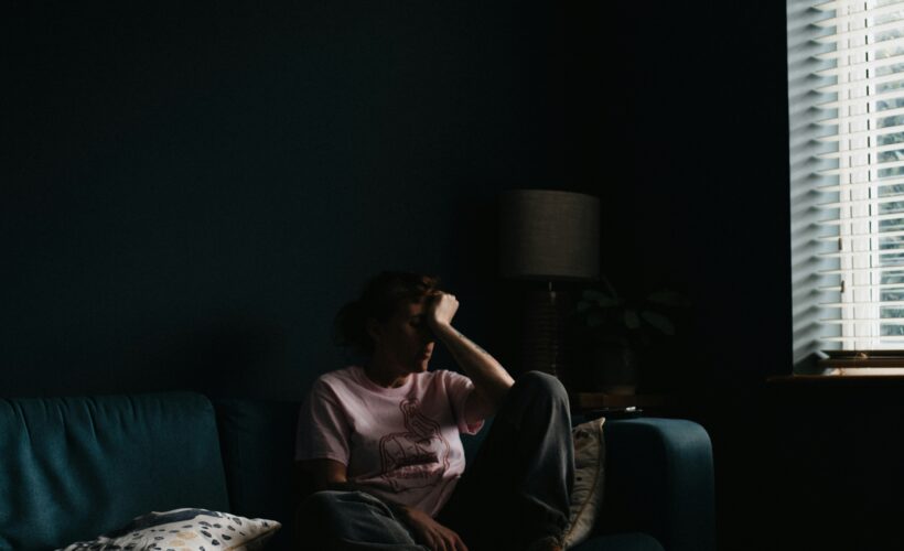 A person is seen sitting on a sofa near a window, they are curled up with their head in their hands and they are in low light to represent Wanting to Feel Seen When You’re Chronically Ill