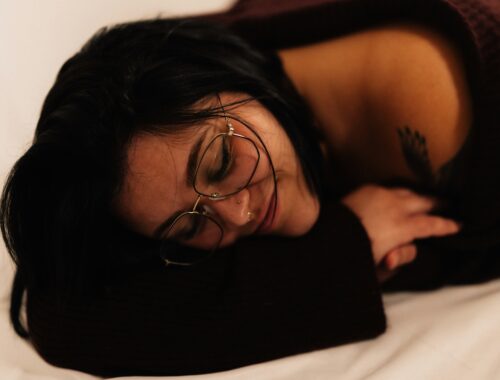 A person is seen curled up in a blanket, their hair is messy and they are wearing glasses, but they look cosy and they are smiling to themselves to represent finding small joys on difficult days.