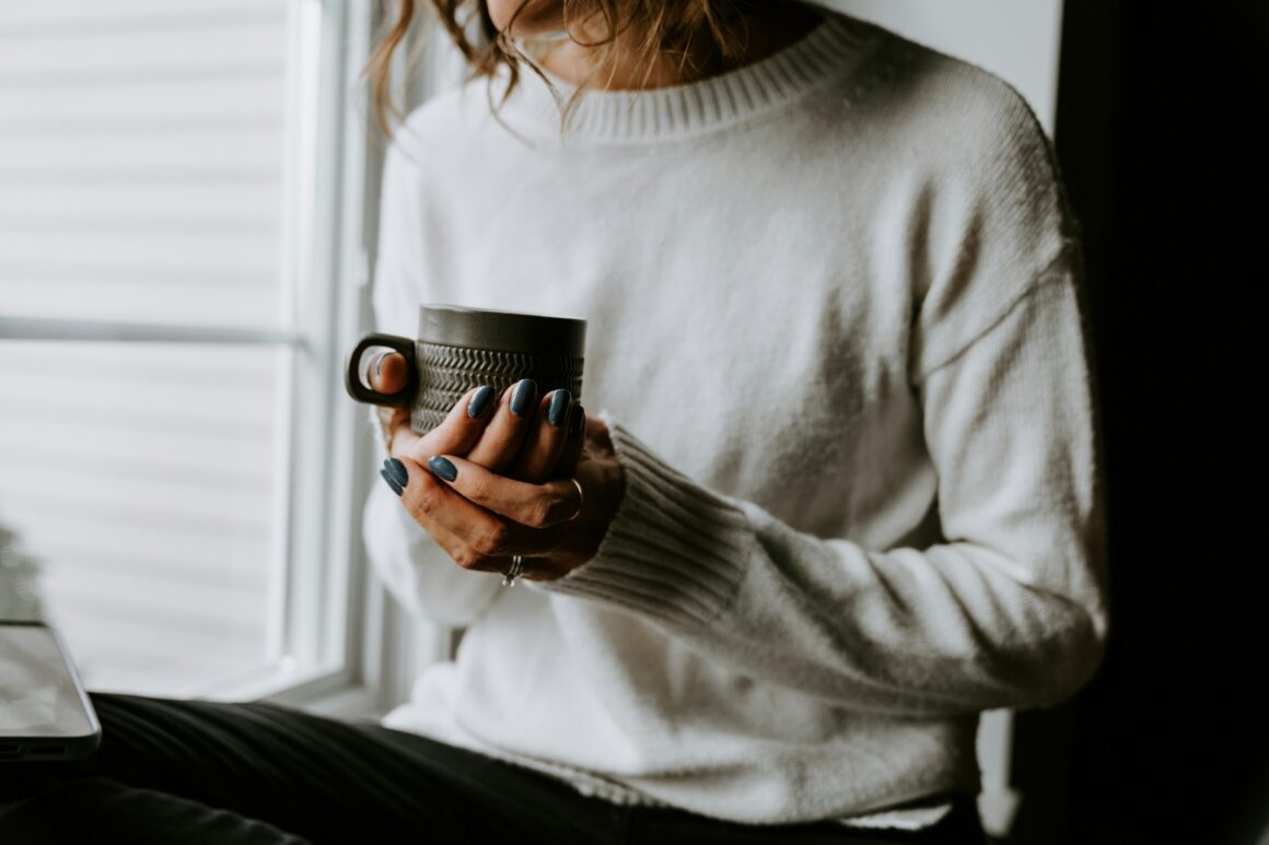 A person is seen sitting in a window, they are wearing a cosy jumper and are holding a mug of hot drink in their hands to represent The Softest Version of Self-Care on Low Energy Days
