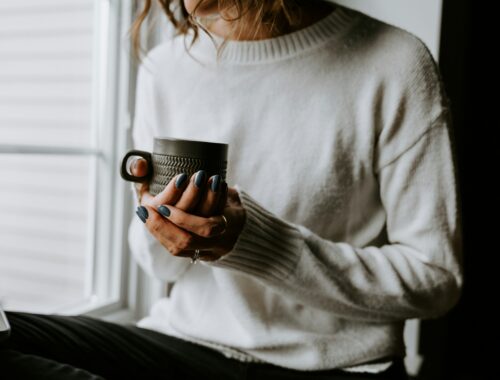A person is seen sitting in a window, they are wearing a cosy jumper and are holding a mug of hot drink in their hands to represent The Softest Version of Self-Care on Low Energy Days