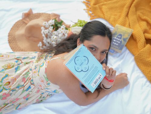 A person is seen on a blanket outside and peering out from behind a book, beside her is a big sunhat and she is wearing a sundress to represent What a Good Day Really Means With Chronic Illness