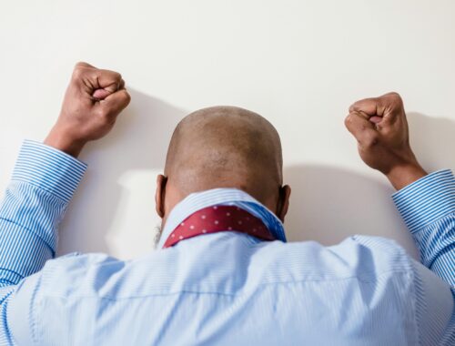 A black man is seen from behind, he is wearing a tie and a blue shirt and is banging his fists on a white wall
