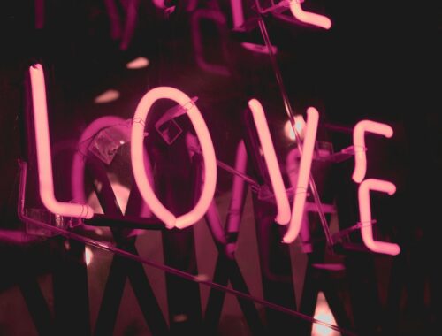 A pink neon sign that says love is seen against a darkened window to represent that you are worthy of love