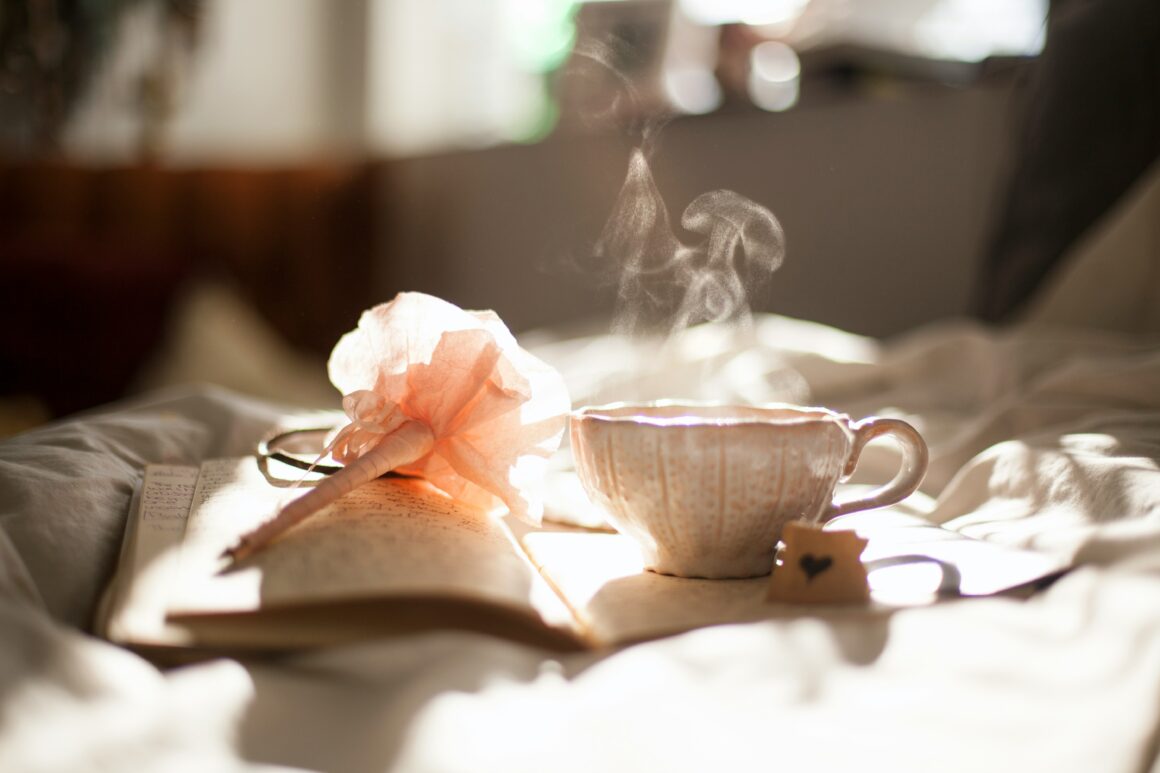 Sunlight falls on an open diary, and a pen with a peach flower on the end, alongside it is a cute teacup with a hand drawn heart on a small card beside it. Steam rises from the cup to represent Finding Joy and Magic While Living with Chronic Illness