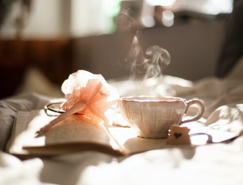 Sunlight falls on an open diary, and a pen with a peach flower on the end, alongside it is a cute teacup with a hand drawn heart on a small card beside it. Steam rises from the cup to represent Finding Joy and Magic While Living with Chronic Illness