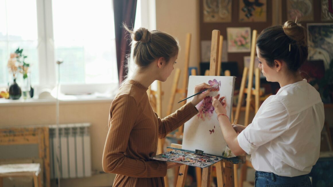Two women paint together in a bright art studio. Female student of art school is painting flowers with oil paints while her teacher good-looking young woman is giving her advice pointing at picture standing near easel.