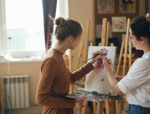 Two women paint together in a bright art studio. Female student of art school is painting flowers with oil paints while her teacher good-looking young woman is giving her advice pointing at picture standing near easel.