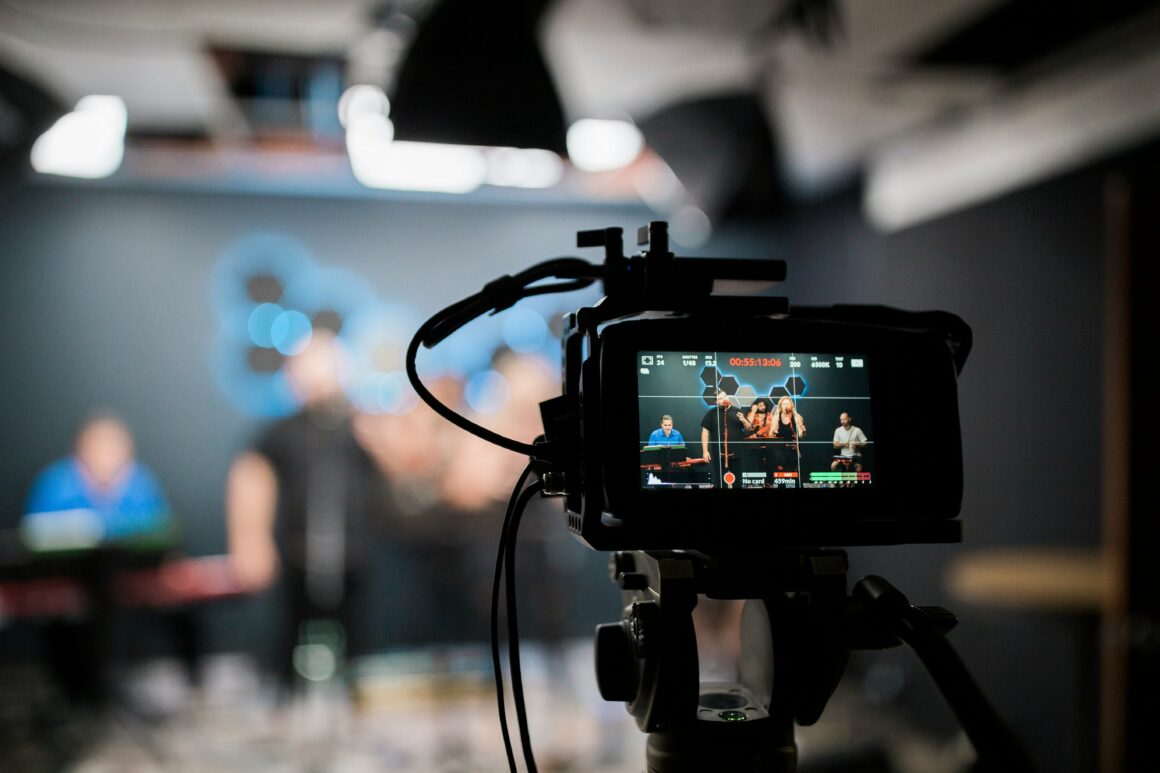 A camera is seen, with behind it blurred, people are recording a video