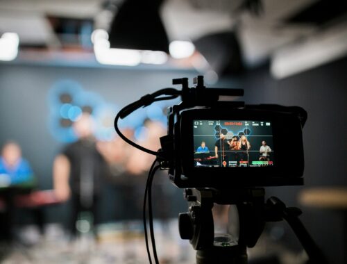 A camera is seen, with behind it blurred, people are recording a video