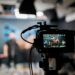 A camera is seen, with behind it blurred, people are recording a video