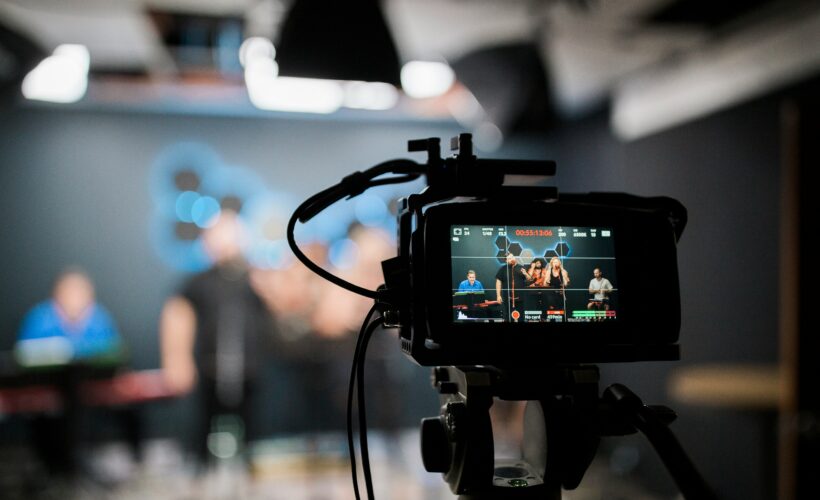 A camera is seen, with behind it blurred, people are recording a video